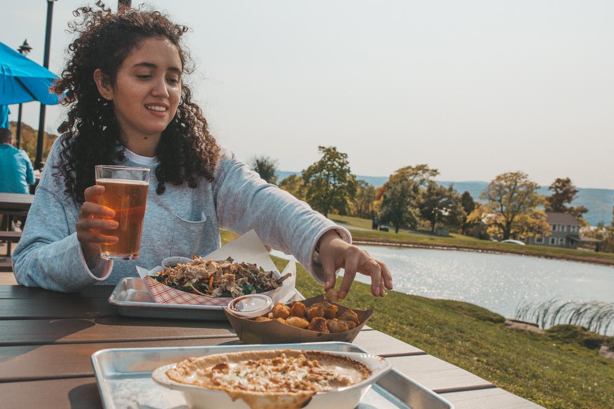 A woman with food and drinks at the outdoor dining area of Grist Iron Brewing Company with a lake in the background